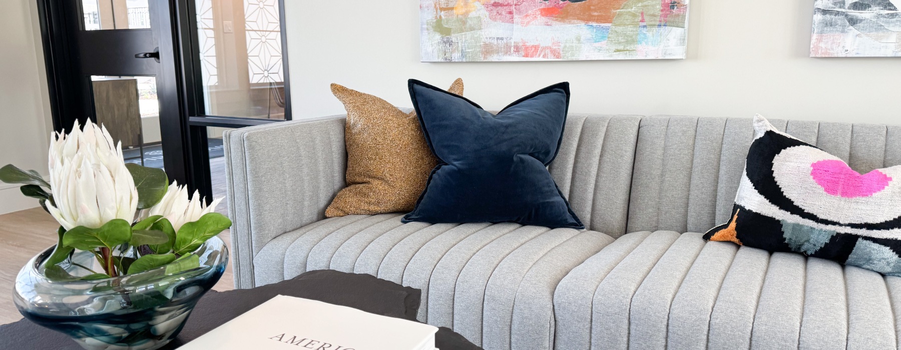 Grey sofa styled with a blue velvet pillow, a multicolor pillow, and a tan pillow, paired with a coffee table topped with a book and a vase.
